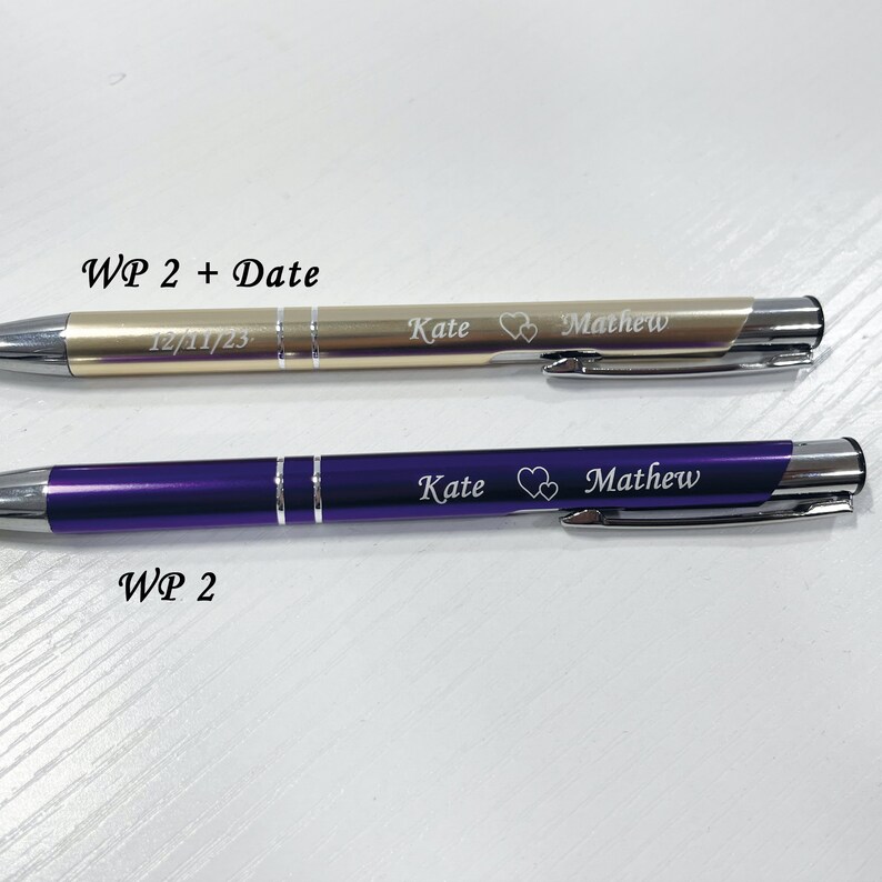 May include: Two pens with personalized engravings. The top pen is gold with the date 12/11/23, the names "Kate" and "Mathew" and a heart symbol. The bottom pen is purple with the names "Kate" and "Mathew" and a heart symbol.