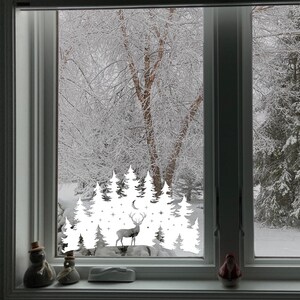 Puede incluir: Adhesivo de vinilo blanco para ventana que presenta una silueta de un ciervo de pie en un bosque nevado con una luna creciente y estrellas. El adhesivo se aplica a una ventana con vista a un paisaje nevado.