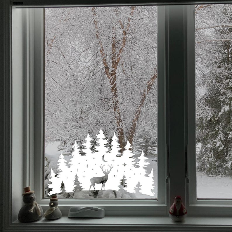 Puede incluir: Adhesivo de vinilo blanco para ventana que presenta una silueta de un ciervo de pie en un bosque nevado con una luna creciente y estrellas. El adhesivo se aplica a una ventana con vista a un paisaje nevado.