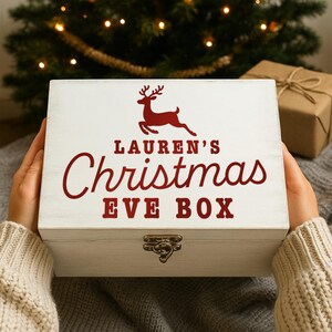 Pu&ograve; includere: Una scatola di Natale in legno bianco con una grafica di renna rossa e la scritta "LAUREN'S Christmas EVE BOX". La scatola ha una chiusura in metallo ed &egrave; tenuta da una persona che indossa un maglione color crema.