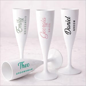 May include: White champagne flutes with personalized names and titles in various colors. The names include "Emily, Georgia, Daniel, and Theo" with titles "Bridesmaid, Bride, Groom, and Groomsman". Ideal for wedding celebrations.