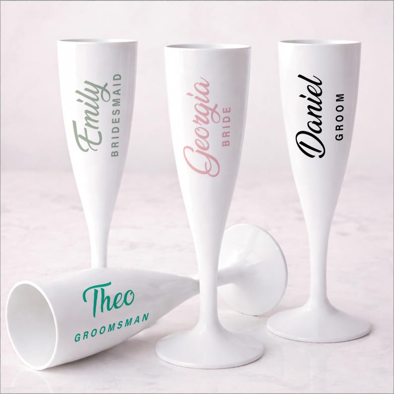 May include: White champagne flutes with personalized names and titles in various colors. The names include "Emily, Georgia, Daniel, and Theo" with titles "Bridesmaid, Bride, Groom, and Groomsman". Ideal for wedding celebrations.