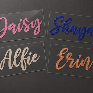 May include: Four clear acrylic name tags with glitter lettering. The names are Daisy in pink, Shayne in blue, Alfie in beige, and Erin in orange. The names are in a cursive font, and the background is dark gray.