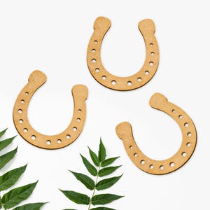 Op de afbeelding: Vier houten hoefijzers met gaten rond de randen, gerangschikt op een witte achtergrond met een groene bladertak in de linkerbenedenhoek.