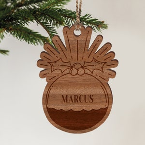 May include: A wooden Christmas ornament shaped like a pudding with a holly sprig and berries on top. The ornament is personalized with the name "MARCUS".