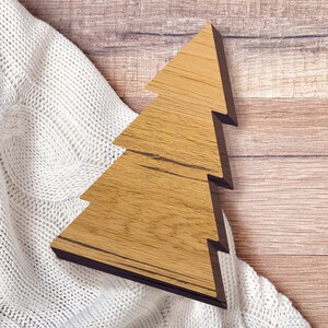 May include: A wooden Christmas tree cutout on a white knitted background. The tree is a light brown color with a dark brown outline.
