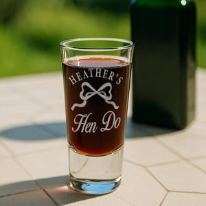 May include: A clear shot glass filled with a dark liquid, likely a spirit or liqueur. The glass is etched with the words "Heather's Hen Do" and a bow design. A dark green bottle is blurred in the background, suggesting a celebratory event.