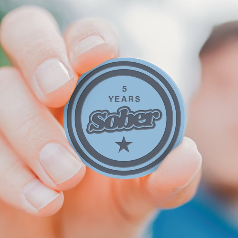 May include: A person's hand holding a blue round token with the text "5 YEARS SOBER" and a star in the center.