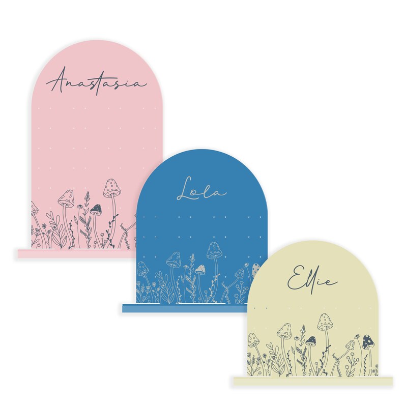 May include: Three pastel-colored arch-shaped signs with names "Anastasia", "Lola", and "Ellie" written in cursive. Each sign has a white outline and a bottom shelf. The signs are decorated with a line of white mushrooms and foliage.