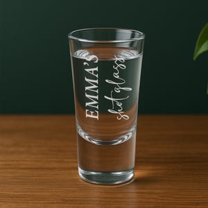 May include: A clear shot glass filled with liquid, featuring the text "EMMA'S shot glass" in a white, elegant script. The glass is cylindrical and sits on a wooden surface, with a dark green background.