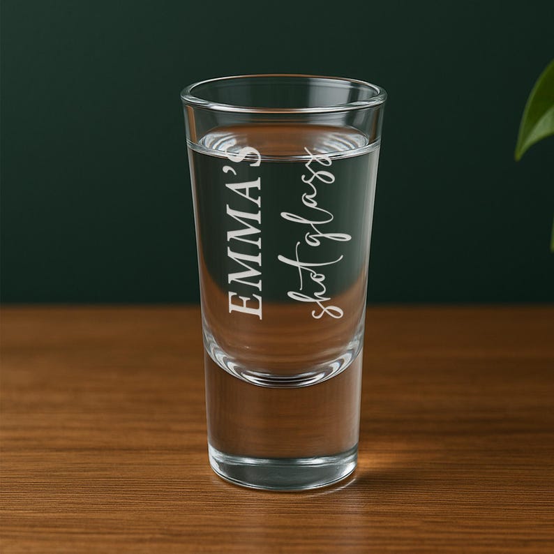 May include: A clear shot glass filled with liquid, featuring the text "EMMA'S shot glass" in a white, elegant script. The glass is cylindrical and sits on a wooden surface, with a dark green background.