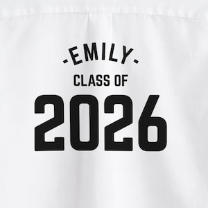 May include: White fabric with the text "-EMILY- CLASS OF 2026" in black. The text is centered and in a bold, sans-serif font. The fabric appears to be a shirt or other garment.