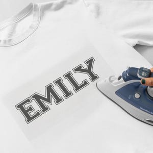 May include: A white t-shirt with the name "EMILY" printed in black letters. An iron is being used to press the letters onto the shirt.