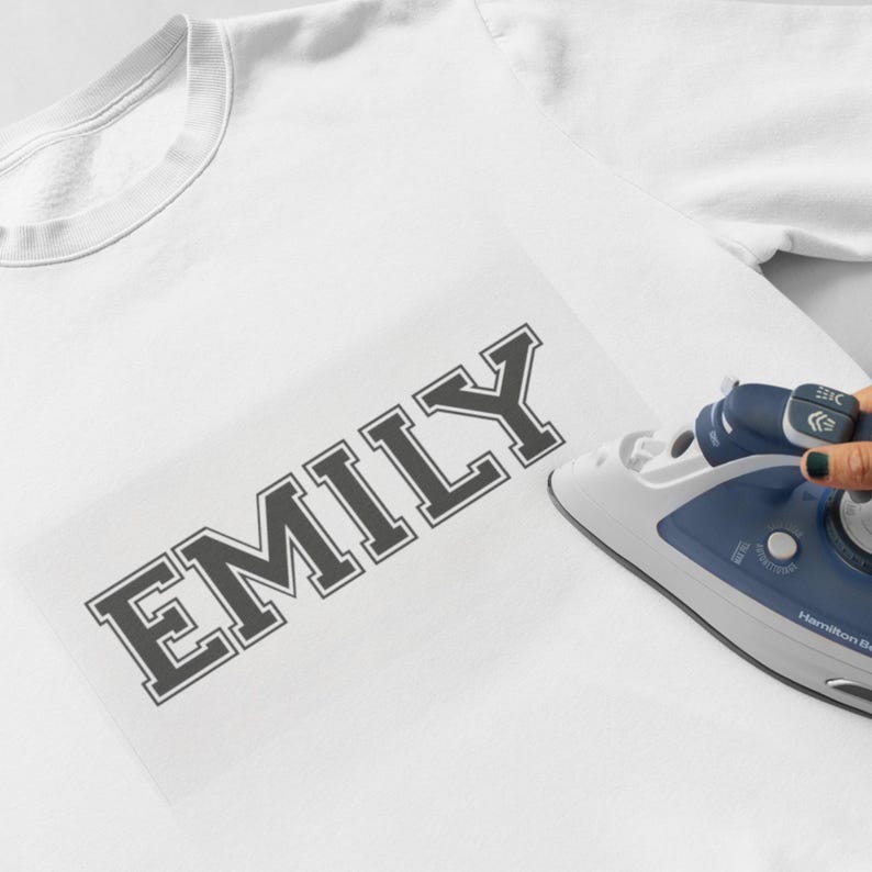 May include: A white t-shirt with the name "EMILY" printed in black letters. An iron is being used to press the letters onto the shirt.