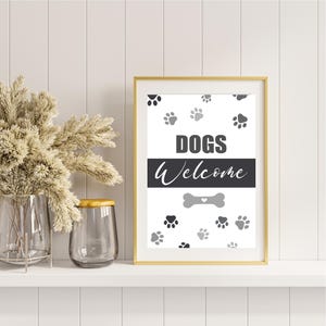 May include: A framed print with a gold border, featuring the words "DOGS Welcome" in black and white. The design includes paw prints and a bone graphic. The print is displayed on a white shelf with decorative items.