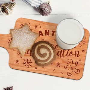 May include: Wooden serving board with the words "Santa's Station" and festive illustrations. A star-shaped cookie, a swirl cookie, and a glass of milk are on the board. Christmas-themed decor is in the background.