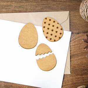 May include: Three wooden Easter egg shapes on a white background. One egg is plain, one has brown polka dots, and one is cracked open.