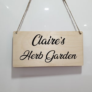 May include: A wooden sign with the text "Claire's Herb Garden" in black cursive lettering. The sign is hanging from a string.