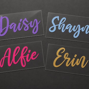 May include: Four clear acrylic name cards with names in various colors. The names are Daisy in purple, Shayne in blue, Alfie in pink, and Erin in gold glitter. The cards are rectangular and sit on a dark surface.
