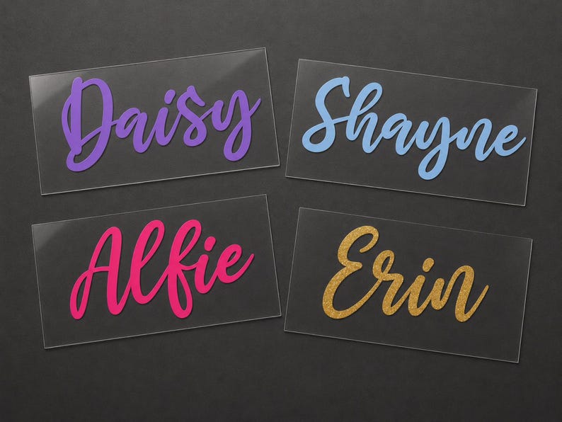 May include: Four clear acrylic name cards with names in various colors. The names are Daisy in purple, Shayne in blue, Alfie in pink, and Erin in gold glitter. The cards are rectangular and sit on a dark surface.