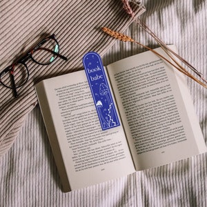 May include: A blue bookmark with white illustrations of mushrooms and flowers. The bookmark has the text "book babe" printed on it.