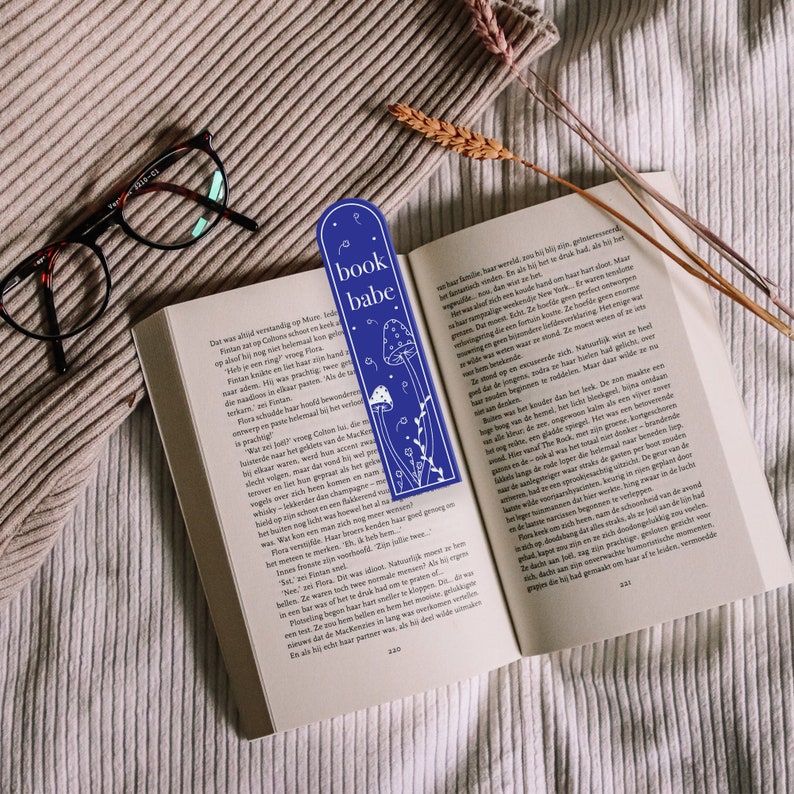 May include: A blue bookmark with white illustrations of mushrooms and flowers. The bookmark has the text "book babe" printed on it.