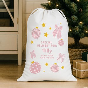 Puede incluir: Saco de regalo navide&ntilde;o blanco con cierre de cord&oacute;n. El saco est&aacute; decorado con adornos, lazos y estrellas rosas. El texto dice: "Special Delivery For: Tilly" y "Do Not Open Until The 25th".