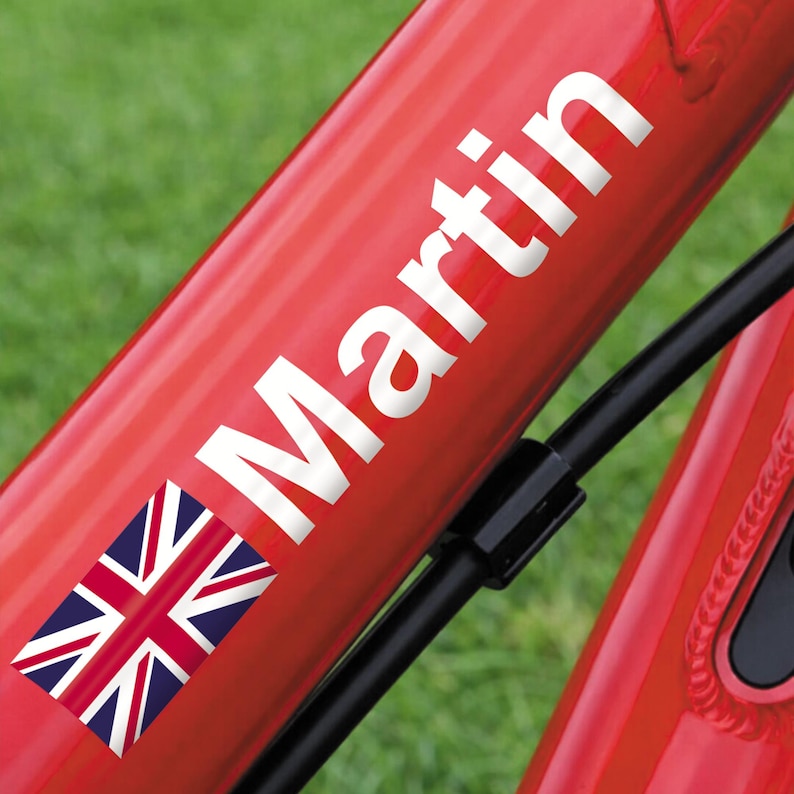 May include: Red bicycle frame with the name "Martin" in white lettering. A Union Jack flag sticker is visible near the name. Black cable and green grass are in the background.