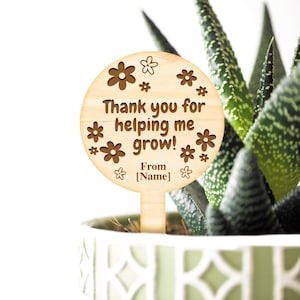 May include: Wooden plant stake with a round, brown sign that says "Thank you for helping me grow! From [Name]" with a flower design.
