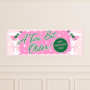 Op de afbeelding: Een roze banner met de tekst "A Tini Bit Older!" in groene letters, en een groene cirkel met "Happy 42nd Birthday Cassie". Martini glazen met olijven staan aan beide uiteinden.