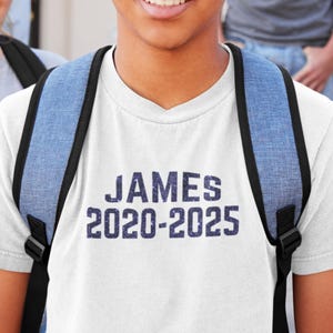 May include: White t-shirt with a blue glitter design that says "JAMES 2020-2025".
