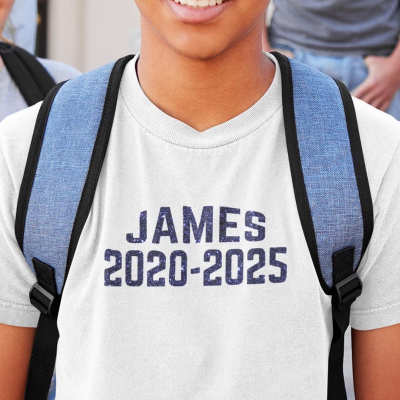 May include: White t-shirt with a blue glitter design that says "JAMES 2020-2025".