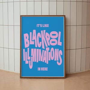 May include: A framed art print with a blue background and pink text that reads "IT'S LIKE BLACKPOOL ILLUMINATIONS IN HERE." The wooden frame surrounds the print, which is displayed against a white tiled wall.