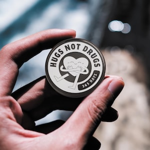 May include: A round, silver pin with a black and white design. The pin features a heart-shaped character with arms and legs hugging itself. The text "Hugs Not Drugs 365 Days" is printed around the character.