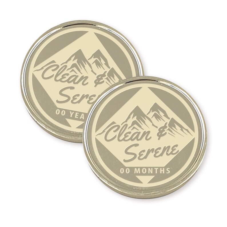 Puede incluir: Dos fichas redondas de color plateado con fondo beige. Las fichas presentan un gr&aacute;fico de una cordillera y el texto "Clean & Serene" con "00 YEARS" y "00 MONTHS" impreso debajo del texto.