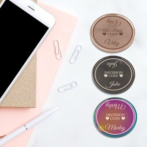 May include: Three round decision coins with different colors and names. The first coin is rose gold with the name "Vicky" engraved on it. The second coin is black with the name "Julie" engraved on it. The third coin is iridescent with the name "Marley" engraved on it.