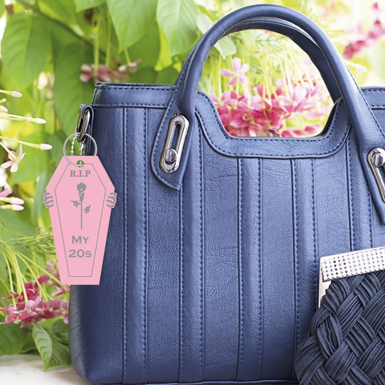 May include: Pink keychain shaped like a coffin with the text "R.I.P. My 20s" and a black rose inside. The keychain is attached to a blue handbag.