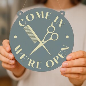 May include: A round, gray sign with the words "COME IN WE'RE OPEN" in a cream color. The sign features a cream-colored comb and scissors graphic. The sign is designed for a hair salon or barber shop.