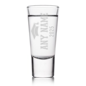 May include: A clear shot glass with the text "ANY NAME 2025" and a graduation cap design etched on the front. The glass is filled with a clear liquid and is set against a white background.