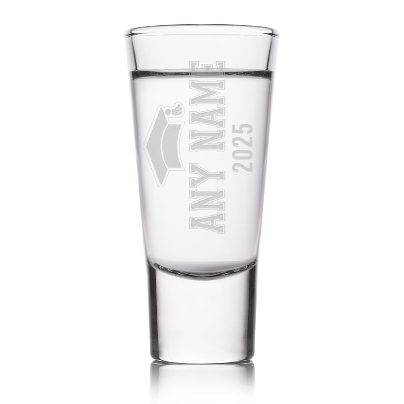 May include: A clear shot glass with the text "ANY NAME 2025" and a graduation cap design etched on the front. The glass is filled with a clear liquid and is set against a white background.