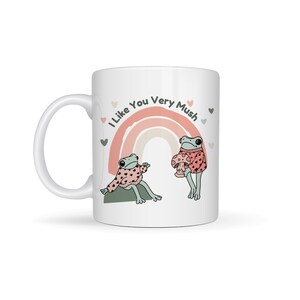 Puede incluir: Taza de cer&aacute;mica blanca con un arco&iacute;ris y dos ranas que llevan jers&eacute;is. El texto "I Like You Very Mush" est&aacute; impreso en la taza.