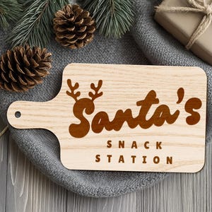 Op de afbeelding: Een lichtgekleurde houten snijplank met een handvat en de woorden "Santa's Snack Station" in bruin ge&euml;tst. Het ontwerp bevat rendiergeweien boven het woord "Santa's".