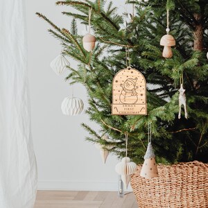 May include: Wooden Christmas bauble featuring a snowman design and the text "Emma's First Christmas". The bauble is hanging on a green Christmas tree.