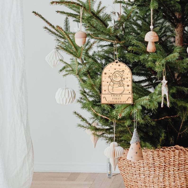 May include: Wooden Christmas bauble featuring a snowman design and the text "Emma's First Christmas". The bauble is hanging on a green Christmas tree.