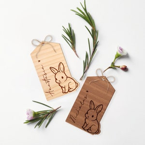 May include: Two wooden gift tags with bunny illustrations and personalized names. The tag on the left is light brown with the name "Anastasia" and the tag on the right is dark brown with the name "Nicholas".