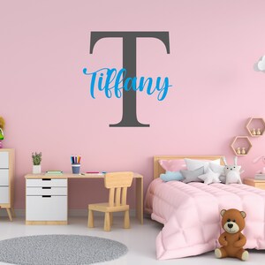 May include: A pink bedroom with a white desk, a wooden chair, and a bed with pink bedding. A large gray letter "T" is on the wall with the name "Tiffany" written in blue cursive.
