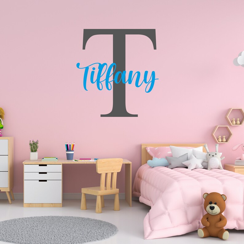 May include: A pink bedroom with a white desk, a wooden chair, and a bed with pink bedding. A large gray letter "T" is on the wall with the name "Tiffany" written in blue cursive.