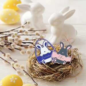May include: Easter-themed decorations featuring two white ceramic bunny figurines, yellow polka-dot eggs, and pussy willow branches. Two egg-shaped name tags with bunny cutouts rest in a nest. One tag is blue with the name "Nicholas", the other is pink with the name "Effie".