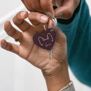 Op de afbeelding: Een donkerbruine hartvormige sleutelhanger met een silhouet van een Franse bulldog en de naam "Guss" in roze schrift.
