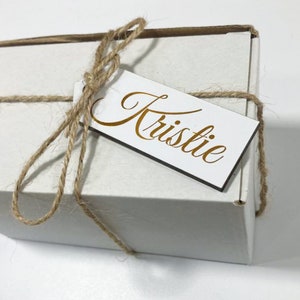 May include: A white gift box tied with brown twine and a white gift tag with the name "Kristie" written in gold lettering.
